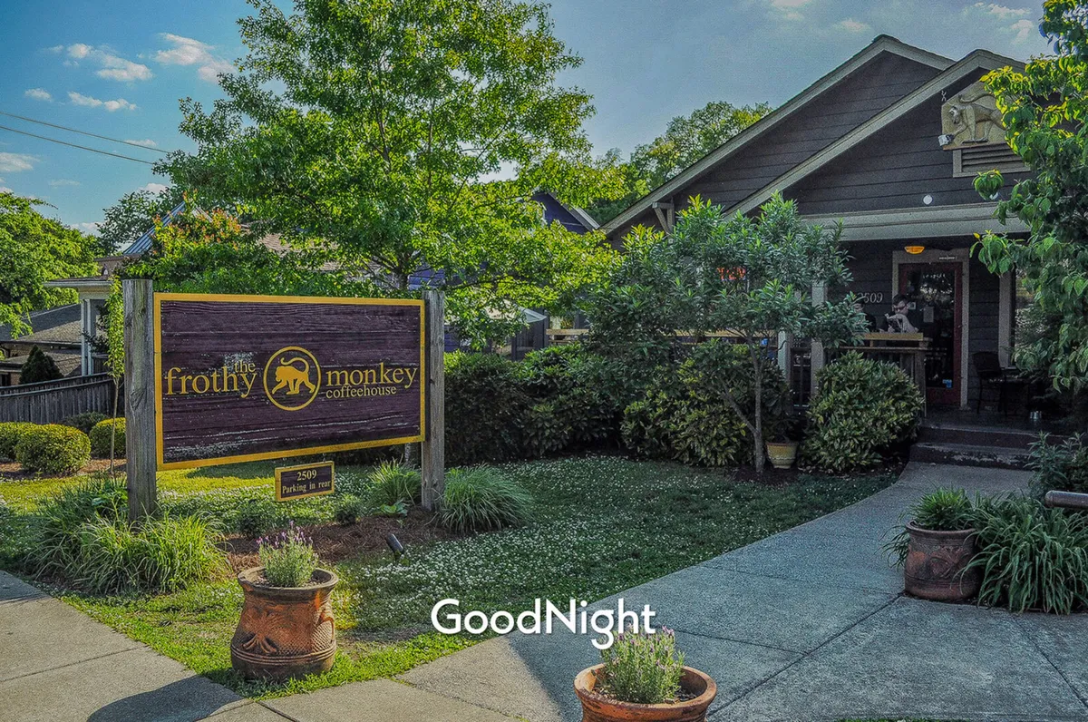 The exterior of 'The Frothy Monkey Coffeehouse' with prominent signage, lush landscaping, and a welcoming entrance. The setting invites passersby to enjoy coffee, socialize, or relax outdoors amidst greenery, suggesting a friendly, community-oriented vibe.