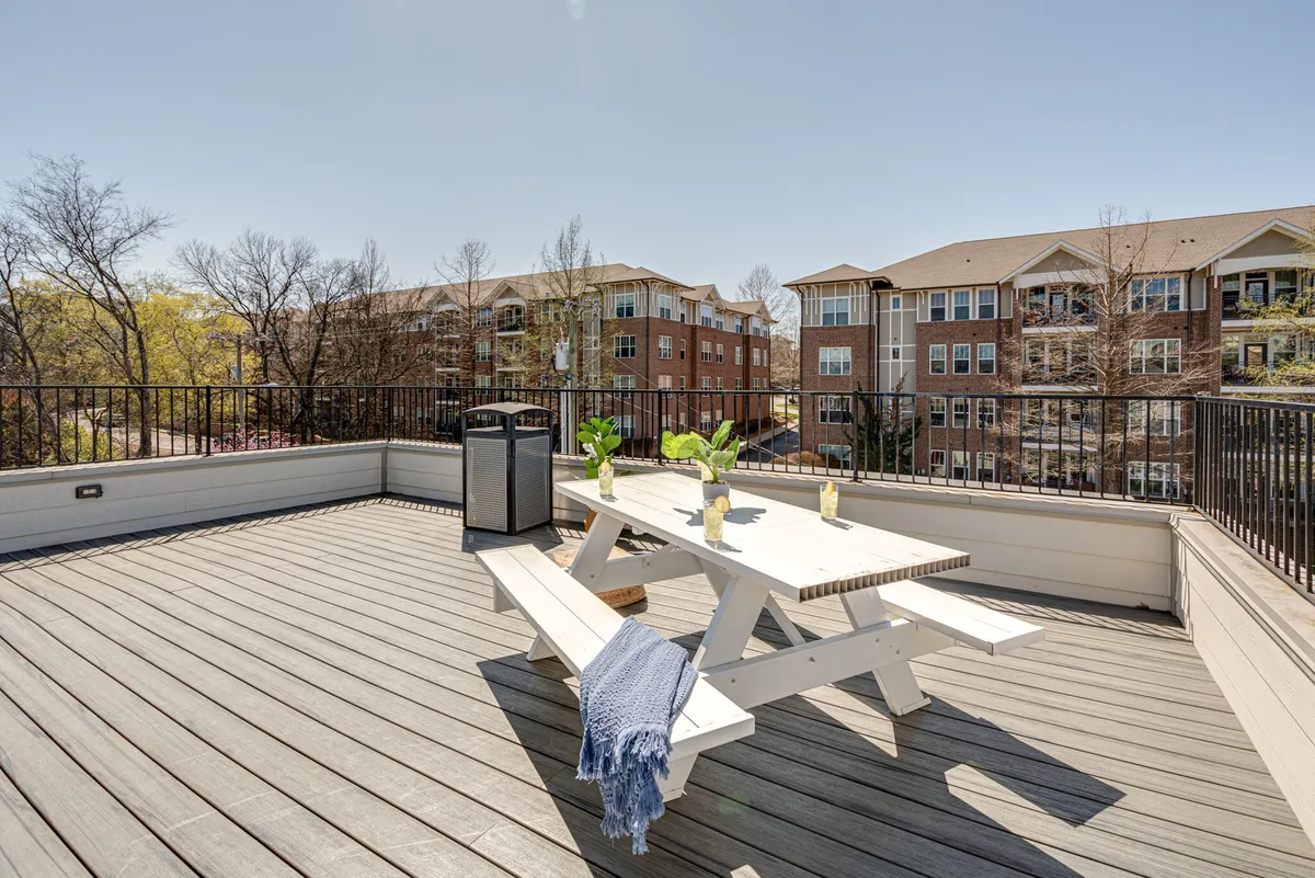 A casual, welcoming outdoor seating area featuring a picnic table surrounded by potted plants and a blue throw blanket, set under the open sky. The scene feels inviting and relaxed, ideal for informal gatherings, meals, or unwinding with friends on the rooftop.
