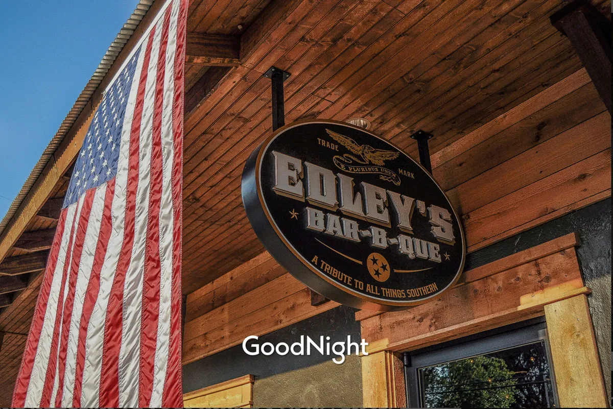 This image features the welcoming exterior of Edley's Bar-B-Que with its distinctive sign and an American flag flying above, suggesting a classic local eatery atmosphere. The setting is inviting, with hints of outdoor activity and a sense of community.