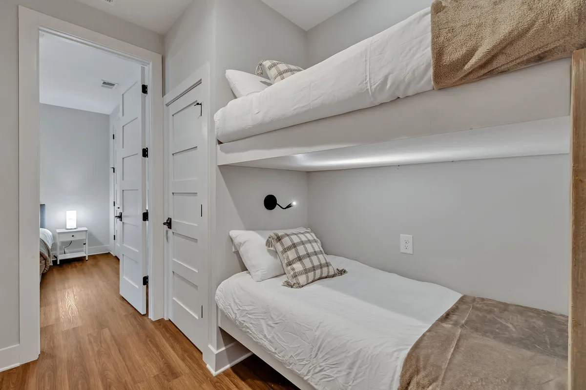 A cozy hallway nook displays twin bunk beds with plaid throw pillows, wood flooring, and a doorway leading to another room. Soft morning light filters in, making the space feel comfortable and perfect for children or friends sharing stories.