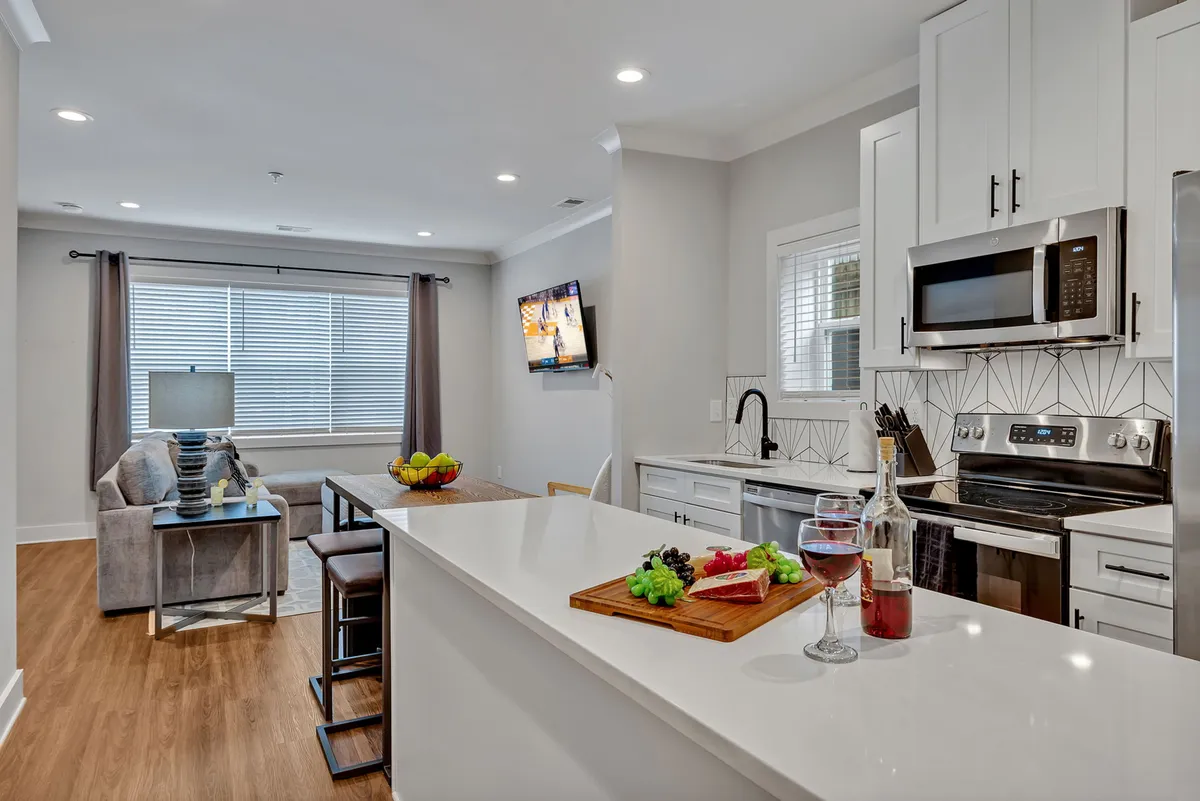 This interior image showcases a welcoming open-plan living area with a view of the kitchen island, living area, large window, and a TV. The space features cozy seating, modern decor, and a layout that encourages movement from the entry into the heart of the home.