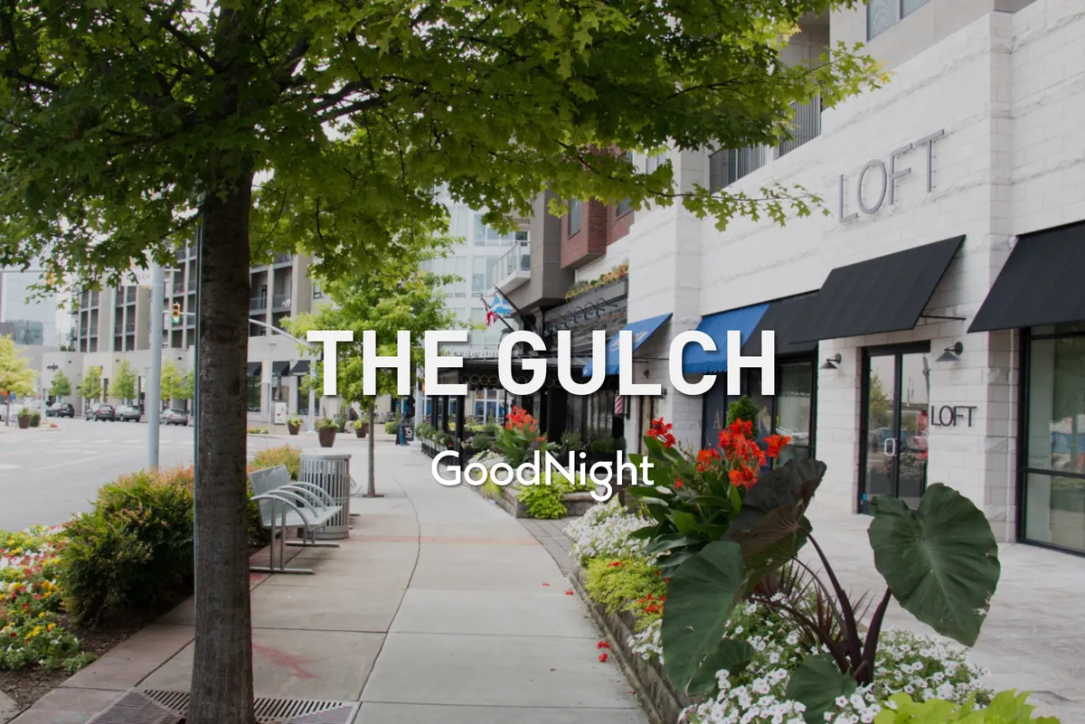 A vibrant exterior streetscape with flourishing green trees, colorful flowers, local storefronts, and benches along the sidewalk. The mood is lively and friendly, with spaces for shopping, relaxing, and socializing, all set beneath a canopy of urban foliage.