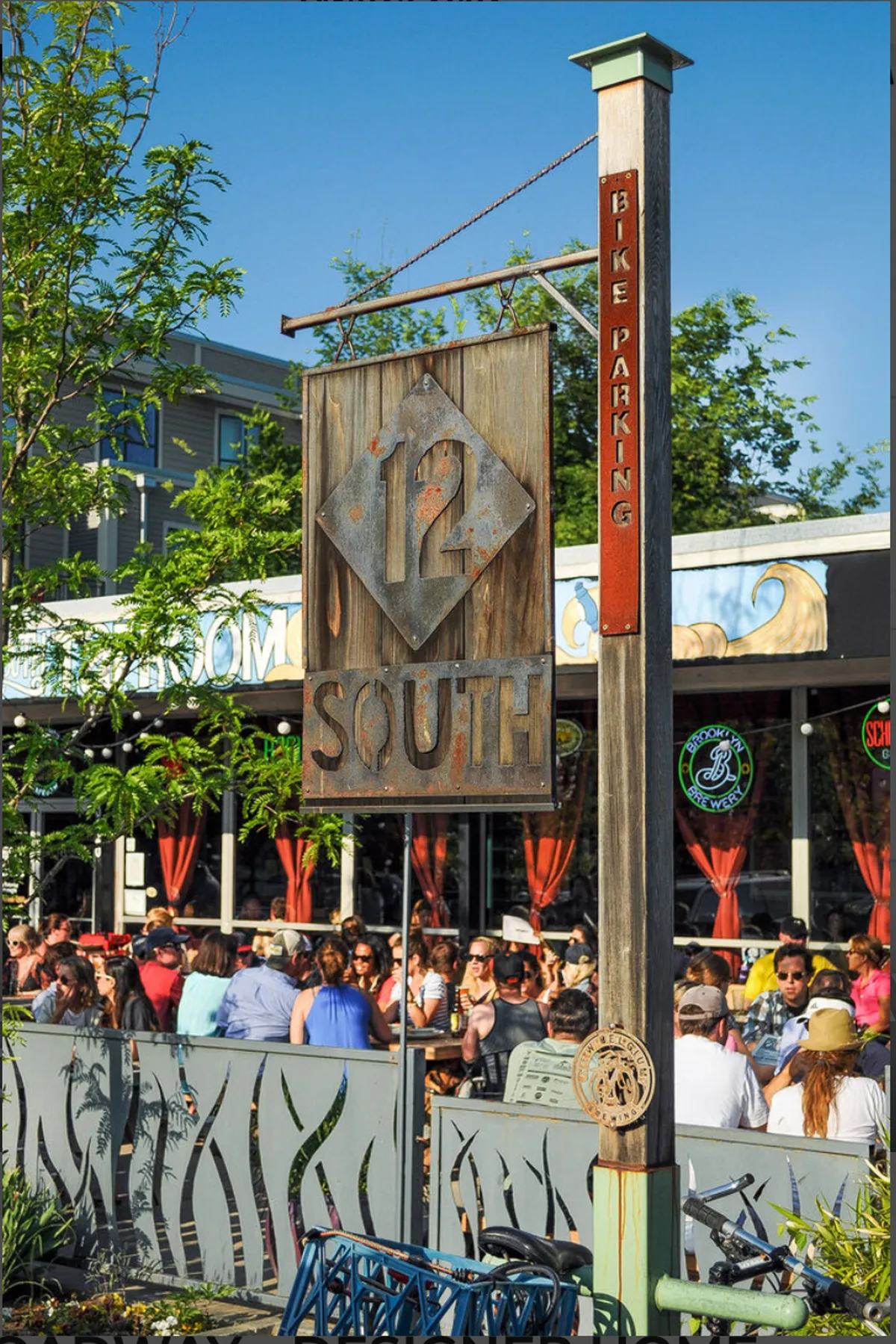 An exterior shot of a bustling street scene featuring the '12 South' sign, bike parking, and an inviting outdoor seating area. People can be seen enjoying meals and socializing, with a friendly, energetic vibe and a strong sense of place.
