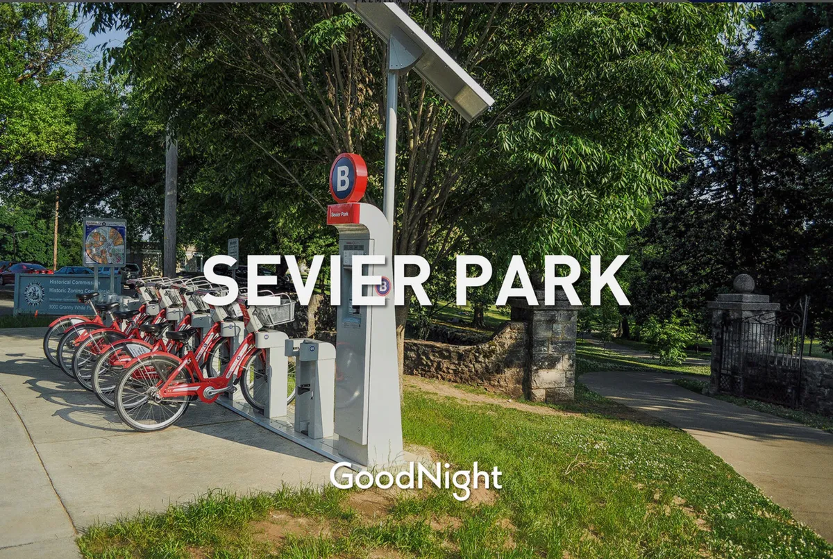 An exterior scene featuring a bicycle rental station positioned beside clear park signage, with abundant greenery in the surrounding area. The image captures an active park environment inviting for biking, walking, picnicking, and community events, with visible open paths and trees that suggest a welcoming outdoor space for pet owners.